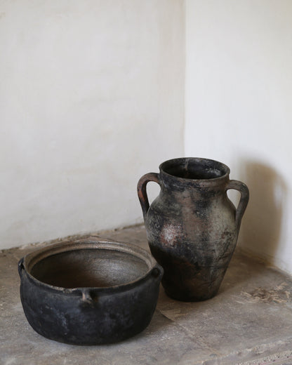 Two ancient-looking black pots on a stone surface with a white wall background