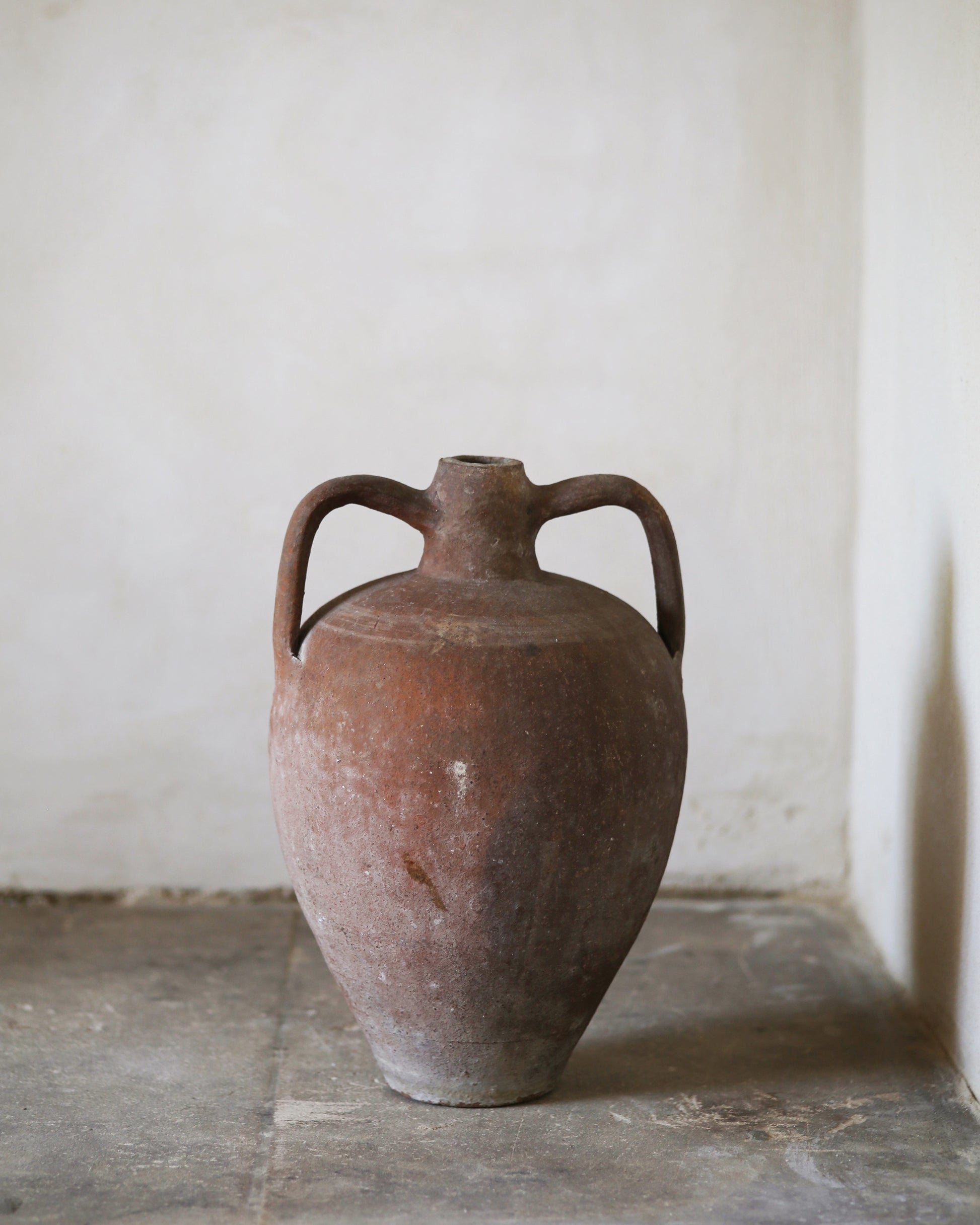 Red terracotta ceramic amphora with handles on a stone surface against a plain wall