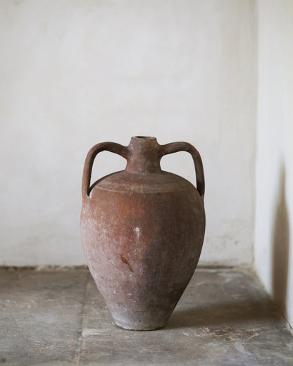 Red terracotta ceramic amphora with handles on a stone surface against a plain wall