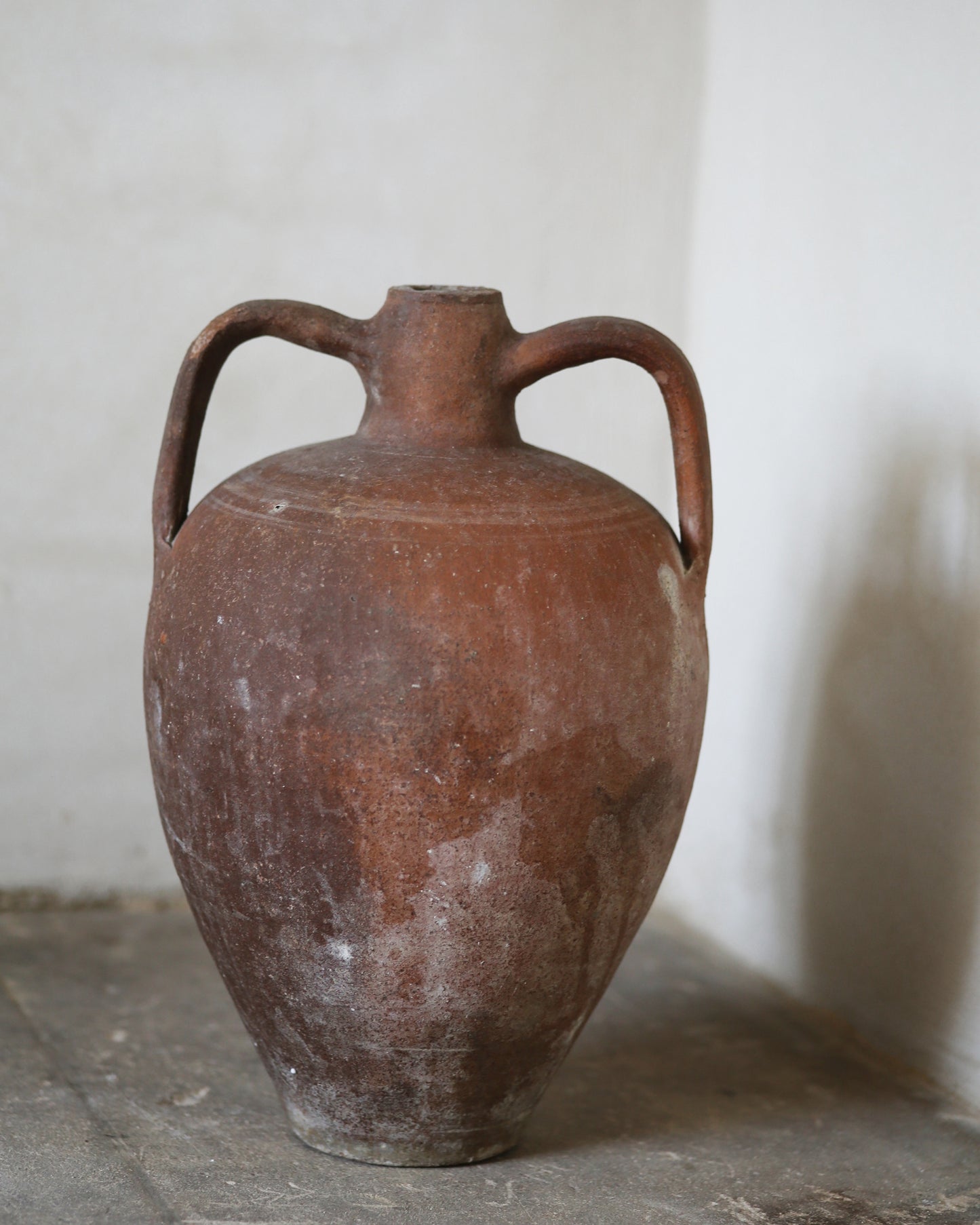 Red terracotta antique amphora with handles on a stone surface against a light background
