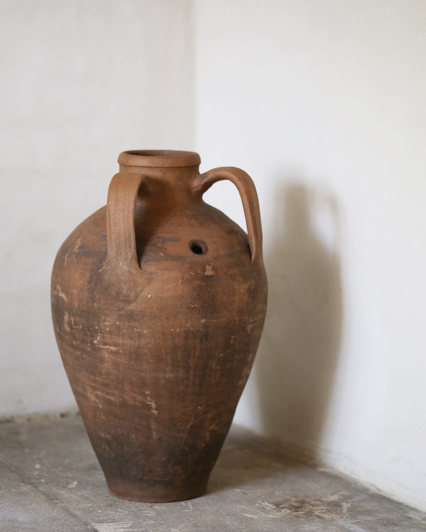 Brown terracotta ceramic jar with handles on flagstone floor against a white wall