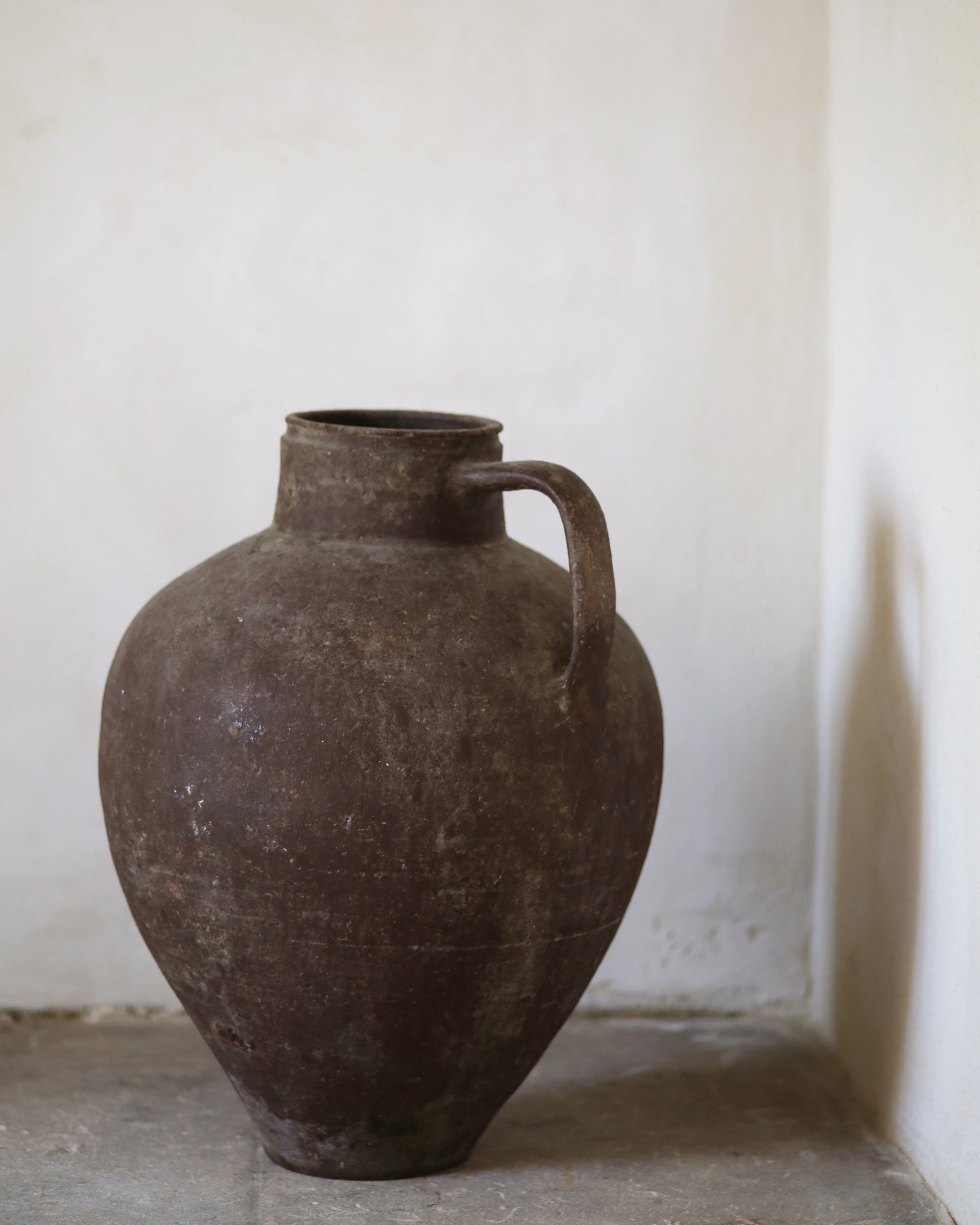 Brown ceramic vase with a handle on a light gray surface against a white wall.