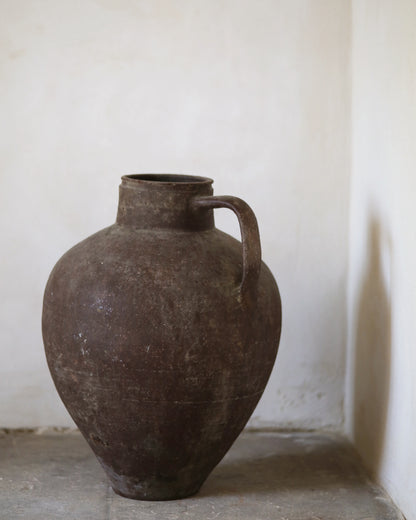 Brown ceramic vase with a handle on a light gray surface against a white wall.