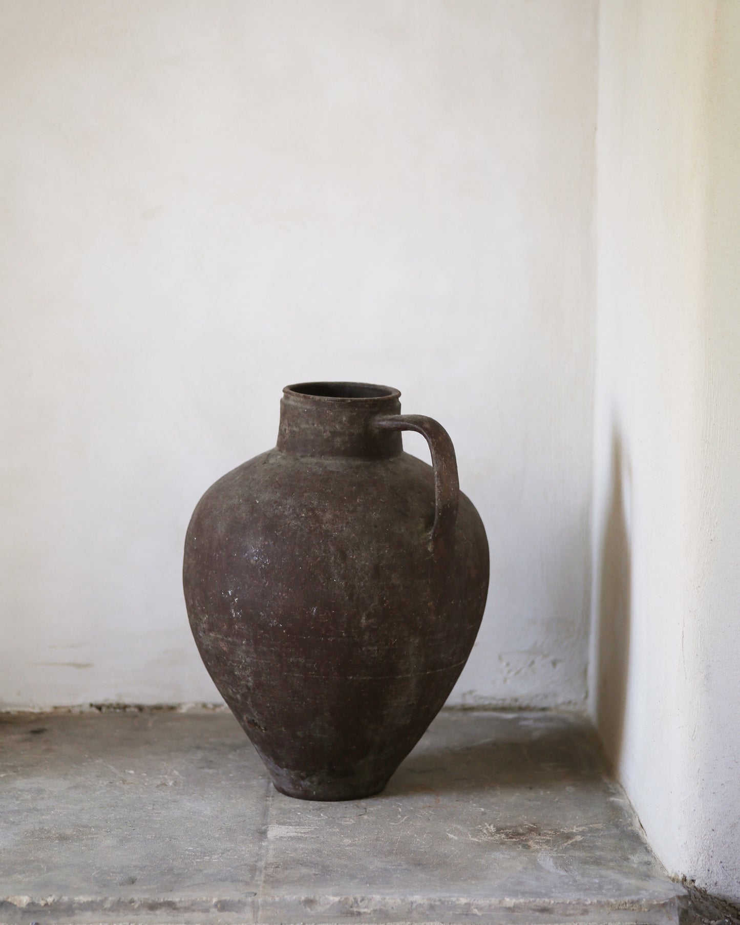 Large brown ceramic jar with a handle on a stone floor against a white wall.