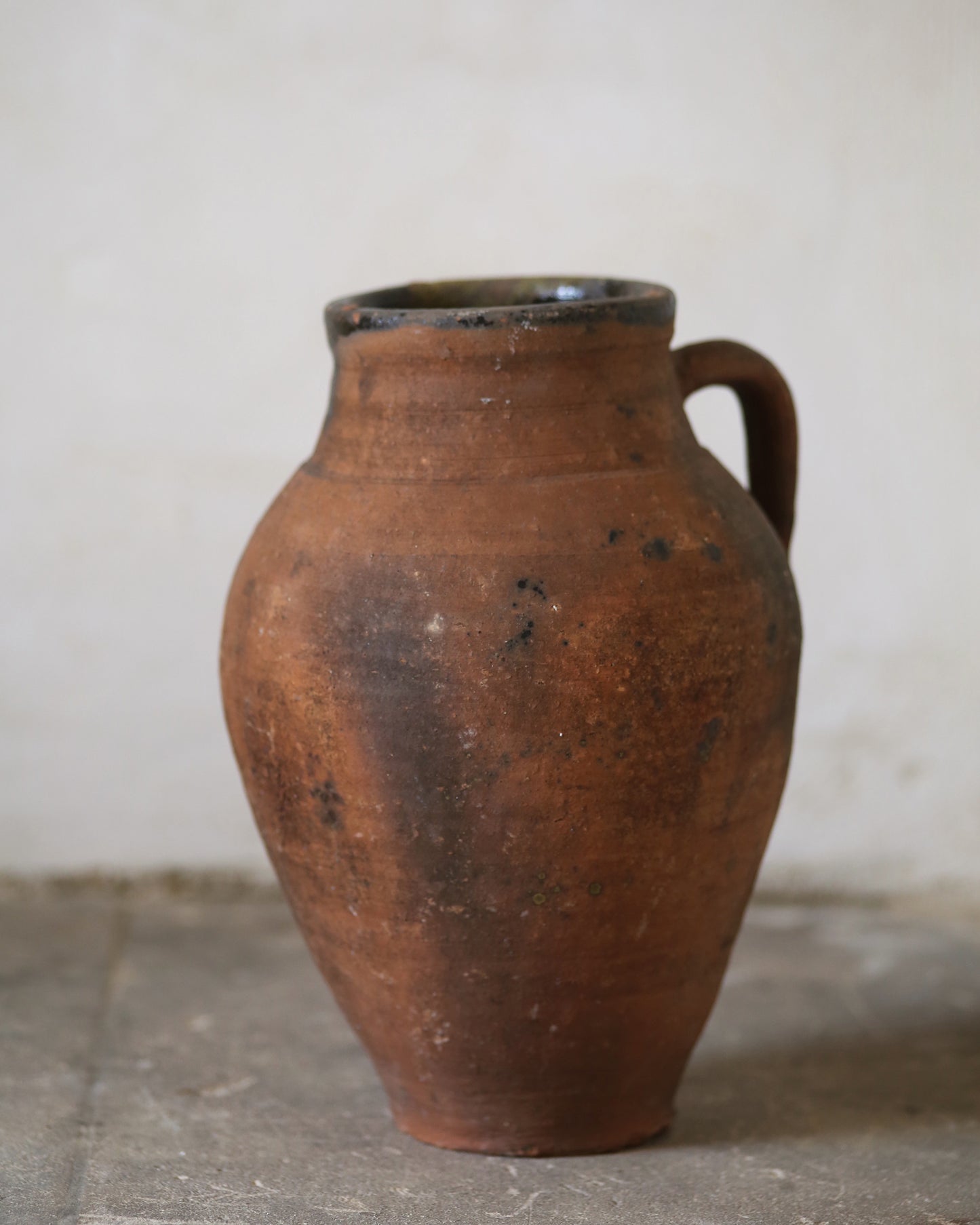 Brown ceramic jar with a handle on a gray floor against a white wall