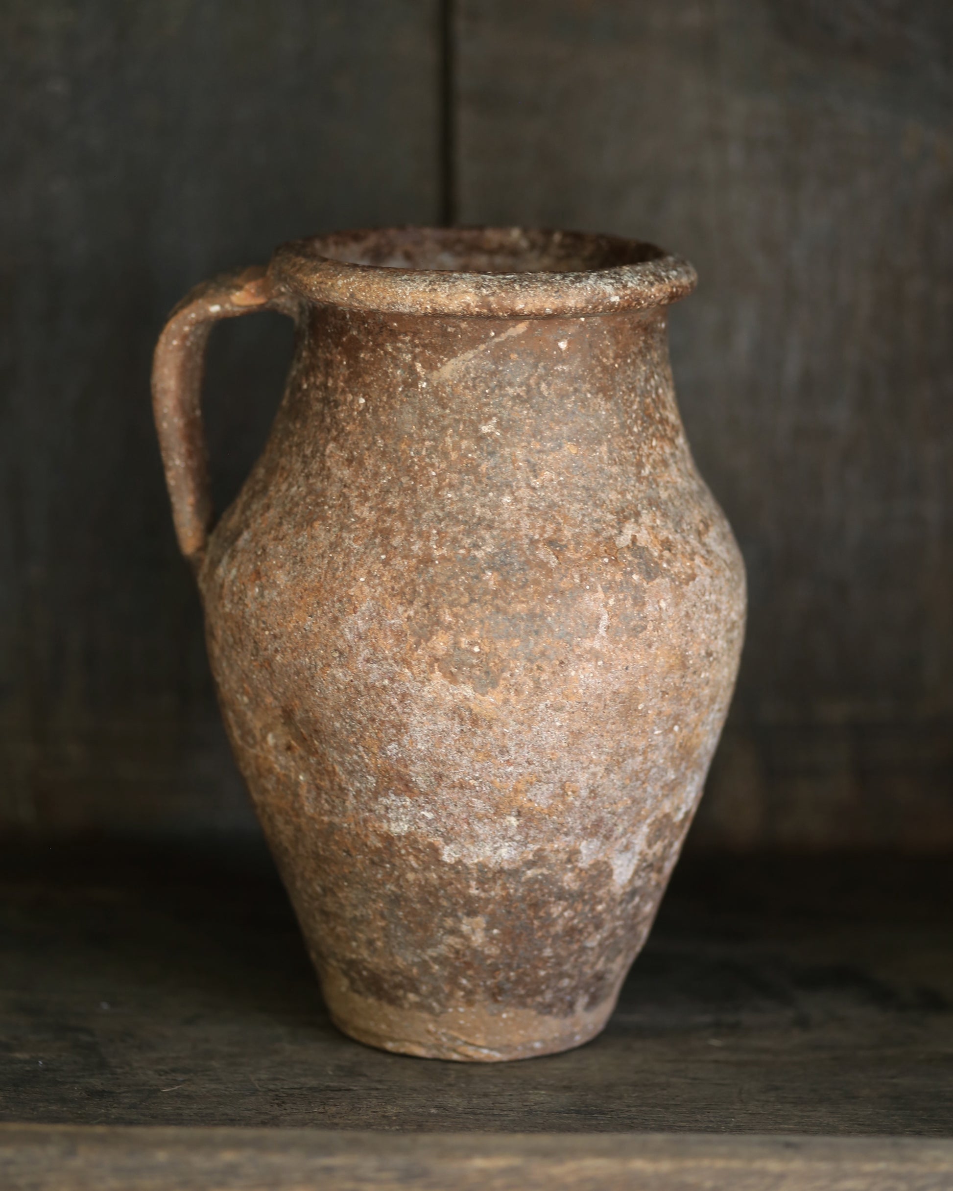 Rustic brown ceramic jar with a handle on a dark shelf