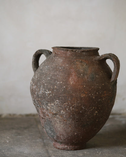 Old antique rustic brown ceramic jar with two handles on a gray surface against a plain background