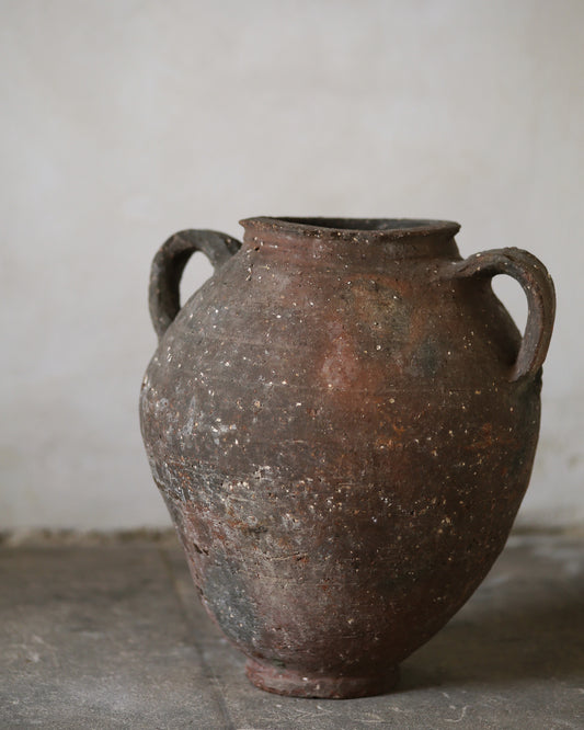 Old antique rustic brown ceramic jar with two handles on a gray surface against a plain background